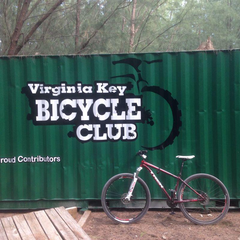 A red mountain bike is leaning against a green shipping container featuring the logo of the Virginia Key Bicycle Club, which reads "Virginia Key BICYCLE CLUB" in bold white letters. The background shows trees, indicating a natural setting. The container is also marked with "Proud Contributors" beneath the main logo. Virginia Key North Point mountain bike trail.