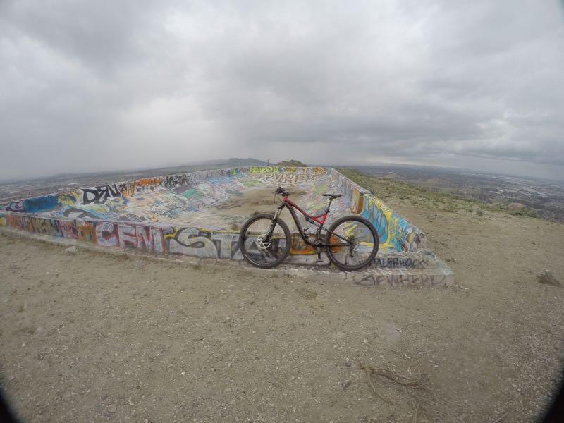 A mountain bike leaning against a graffiti-covered concrete structure on a rocky hilltop, with a cloudy sky and distant views of the landscape below. Battle Bowl mountain bike trail.