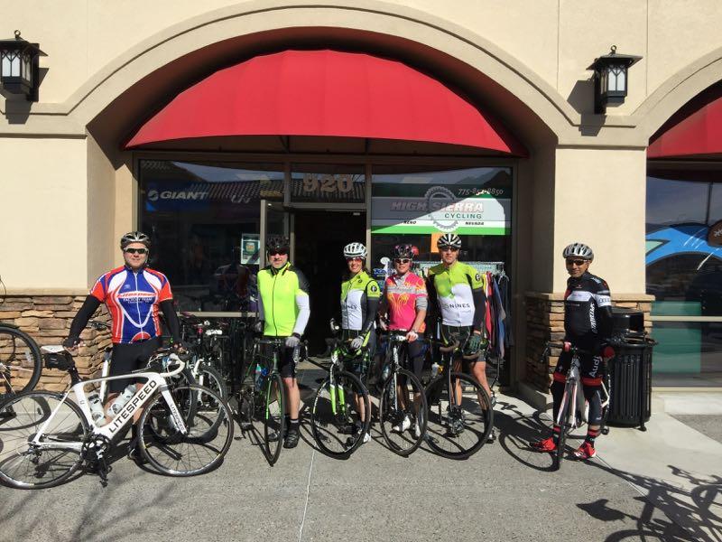 A group of six cyclists, wearing colorful cycling gear, pose with their bikes in front of a bike shop. The setting features a building with a red awning and a large window displaying cycling equipment. The cyclists are smiling and appear ready for a ride, with two bikes prominently displayed in the foreground.