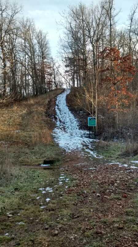 A snowy hiking trail leading uphill through a forest, with bare trees on either side and a signpost visible on the right side of the path. The ground is a mix of grass and mud, with patches of snow scattered along the trail. Slaughter Pen Trail mountain bike trail.