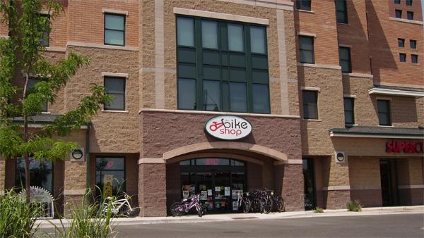 A storefront of a bike shop featuring large windows, a prominent circular sign with the text "bike shop," and a selection of bicycles displayed outside. The building has a mix of materials including brick and stone, set in a neighborhood with residential and retail spaces.