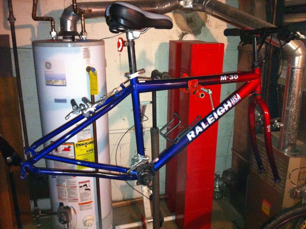 Raleigh Hard tail Mountain Bike: A partially assembled mountain bike in a basement setting, featuring a red and blue frame with the brand name "Raleigh USA" prominently displayed. The bike hangs from a hook, with various components such as the saddle and handlebars visible, alongside a water heater and other household items in the background.