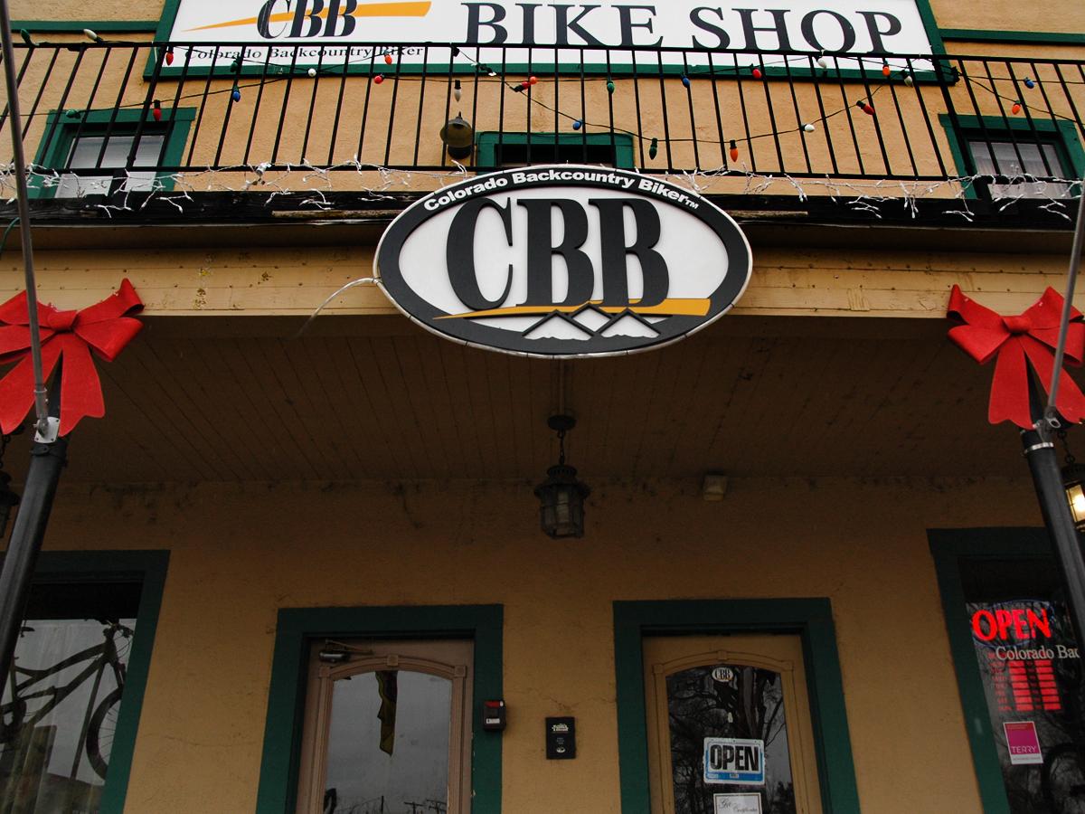 Alt text: Entrance of the Colorado Backcountry Biker bike shop, featuring a large circular sign with the initials "CBB" and decorative bows hanging on either side. The shop has an "OPEN" sign displayed by the door.