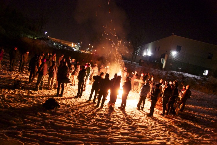 A group of people gathered around a large bonfire on a snowy terrain at night, with sparks and smoke rising from the flames. In the background, there are buildings illuminated by artificial lights. The scene captures a social gathering, with attendees wearing winter clothing and some holding drinks or items.