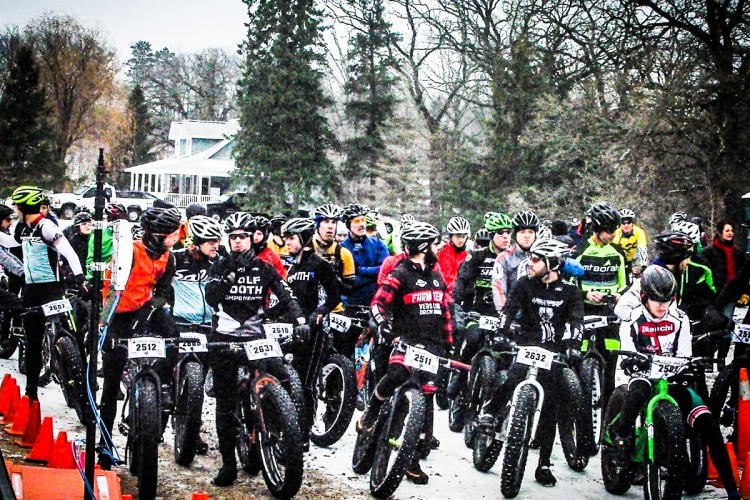 A group of cyclists wearing helmets and various biking gear gathers at the starting line of a race, with snowflakes falling around them. They are lined up next to their fat bikes, and colorful cones mark the course. Sparse trees and a house are visible in the background.