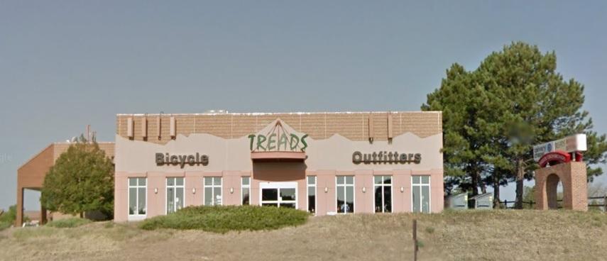 Front view of a building with large windows and a sign that reads "Treads" above the entrance, flanked by the words "Bicycles" and "Outfitters." The exterior is light-colored, and there are green bushes and trees in front. A sign structure can be seen to the right of the building. The sky is clear with no clouds.