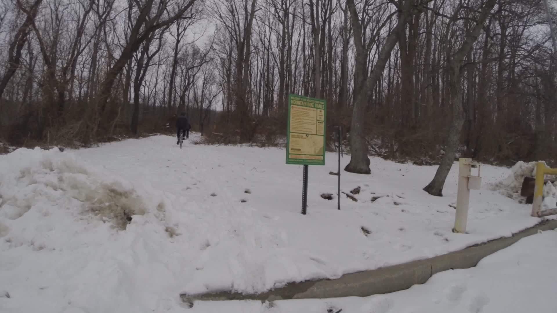 A snowy trail in a forest with leafless trees on either side. A green informational sign about mountain bike trails is visible in the foreground, while a person riding a bike can be seen in the distance traveling along the snowy path. The ground is covered with a thick layer of snow, suggesting winter conditions. Wolfes Pond park mountain bike trail.