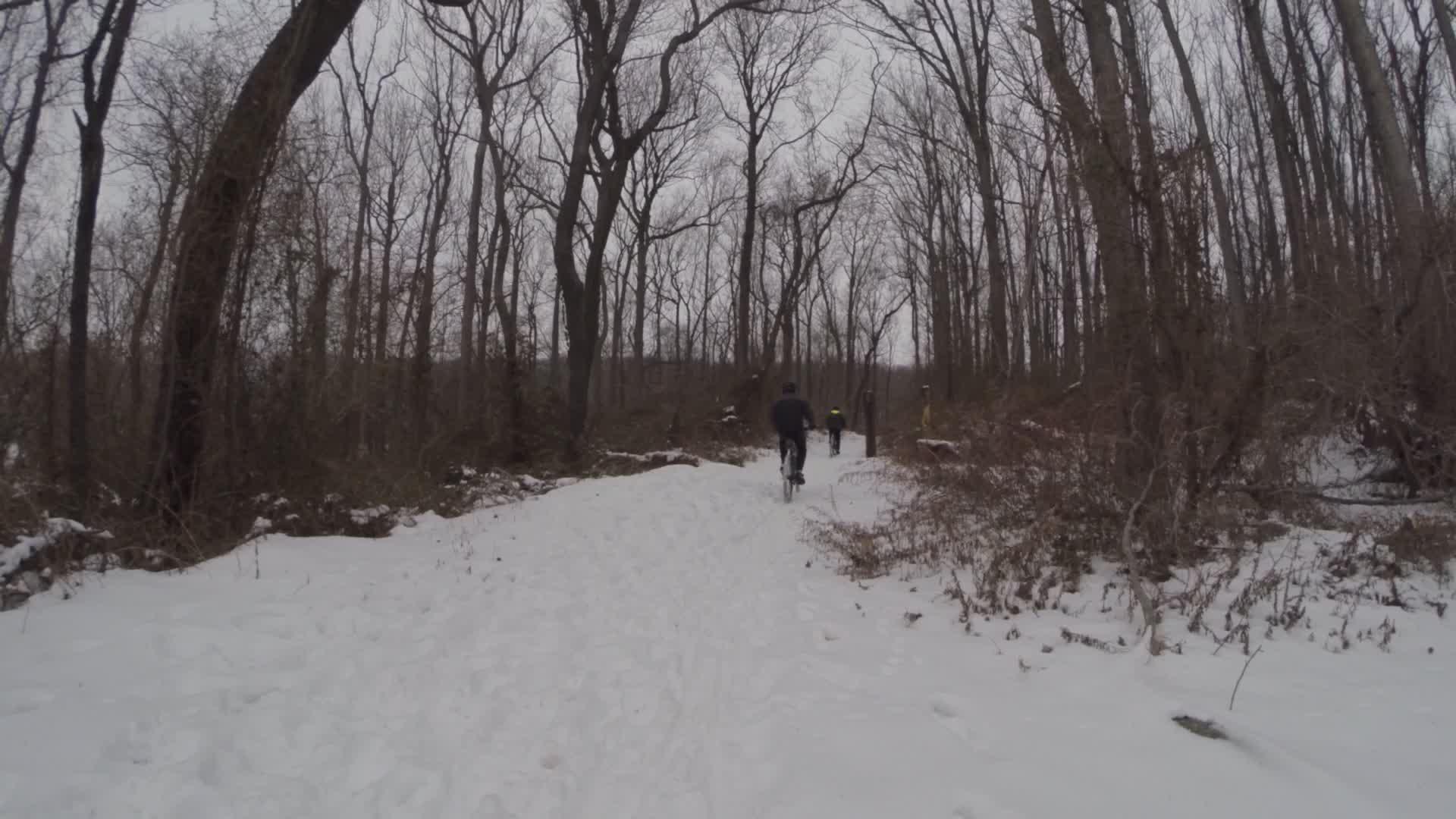 Two cyclists riding on a snow-covered path through a wooded area with bare trees and a cloudy sky. The trail is partly obscured by snow, and there are footprints visible in the snow. Wolfes Pond park mountain bike trail.