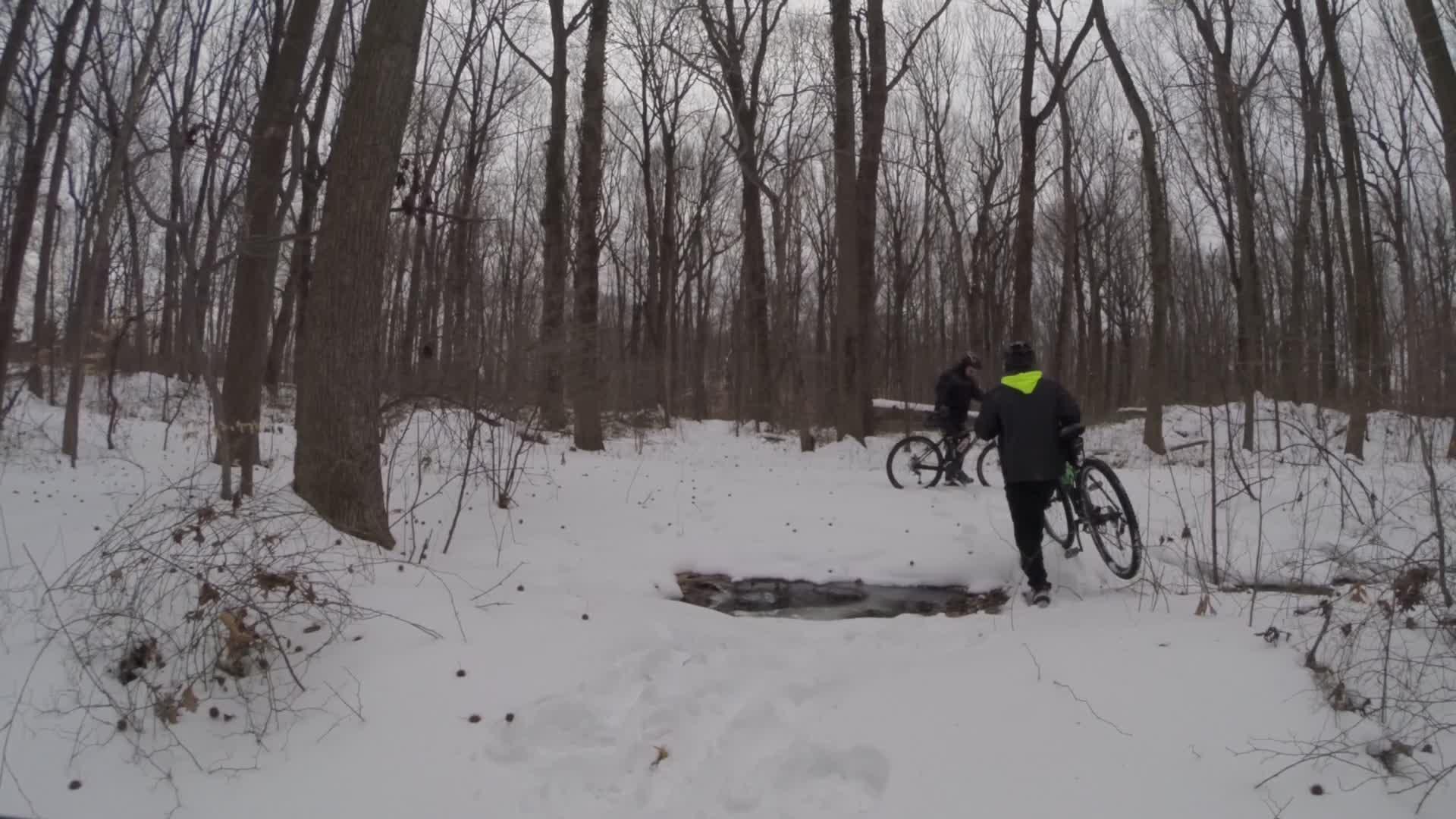 Two mountain bikers navigating a snowy trail in a wooded area, with bare trees in the background and a small stream visible. One biker is carrying their bike while the other is preparing to cross the stream. Trails seperated by streets mountain bike trail.
