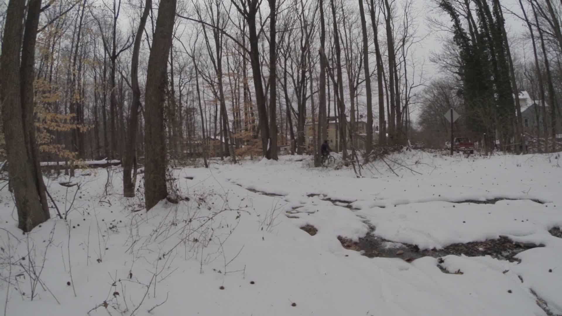 A snowy landscape in a wooded area, featuring tall, barren trees and a small creek running through the snow-covered ground. In the background, a few residential buildings are partially visible, and a person can be seen biking along a snow-covered path. The scene is muted and overcast, typical of winter weather. Trails seperated by streets mountain bike trail.