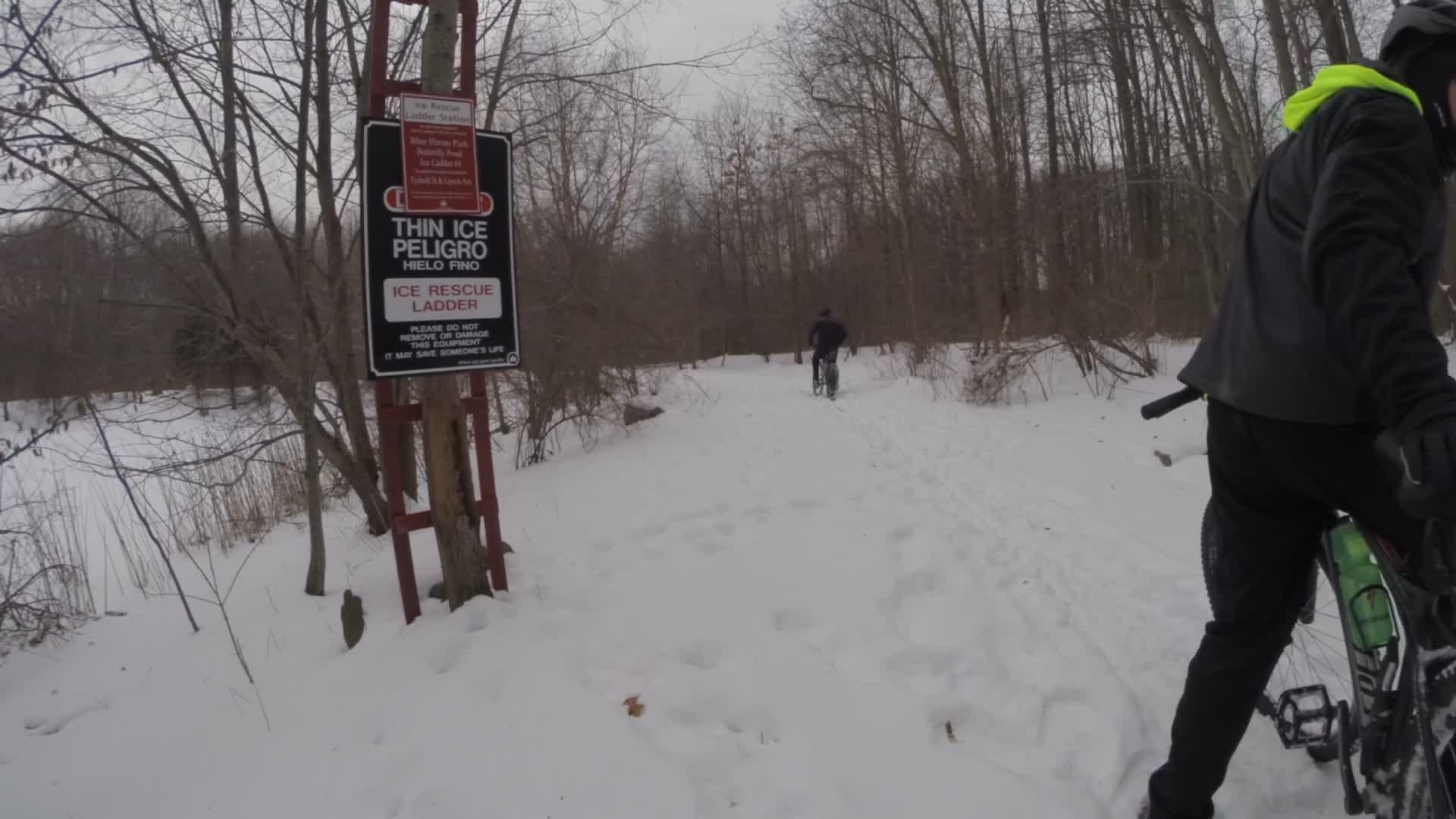A snow-covered path in a wooded area, featuring a warning sign about thin ice and an ice rescue ladder. Two individuals, one riding a bike and another walking, are making their way down the trail. The landscape is bare, typical of winter, with patches of snow and leafless trees surrounding them. Trails seperated by streets mountain bike trail.