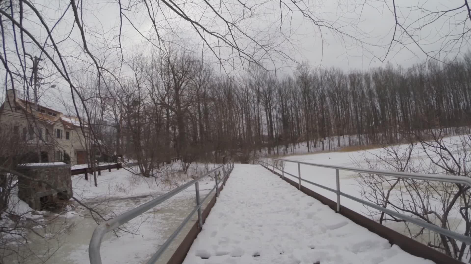 A snow-covered bridge leading over a frozen river, surrounded by bare trees and a cloudy sky. A house is visible on the left, partially obscured by branches. The scene conveys a tranquil winter landscape. Trails seperated by streets mountain bike trail.