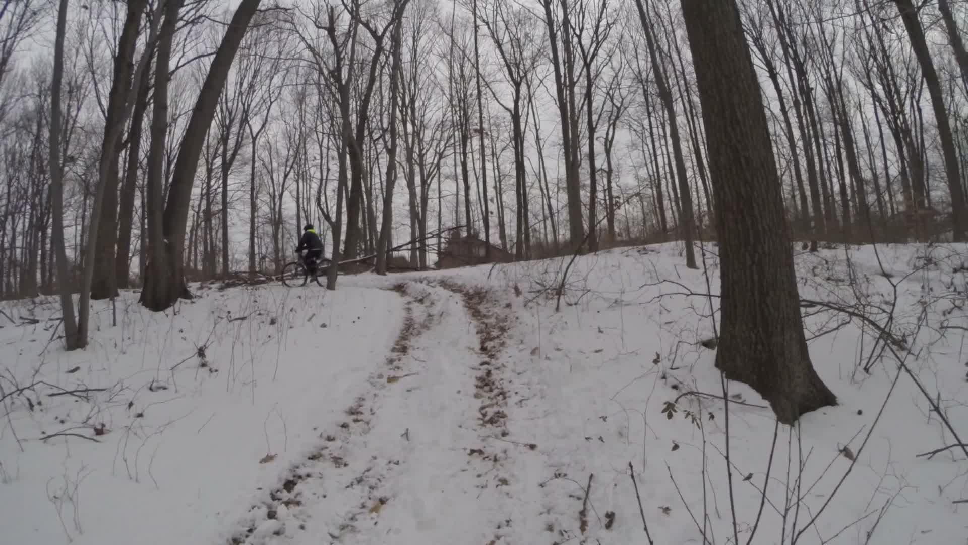 A cyclist riding a mountain bike on a snow-covered trail winding through a forest. The trees are bare, indicating winter, and a light layer of snow blankets the ground. The path shows tire tracks leading through the snowy landscape. Trails seperated by streets mountain bike trail.