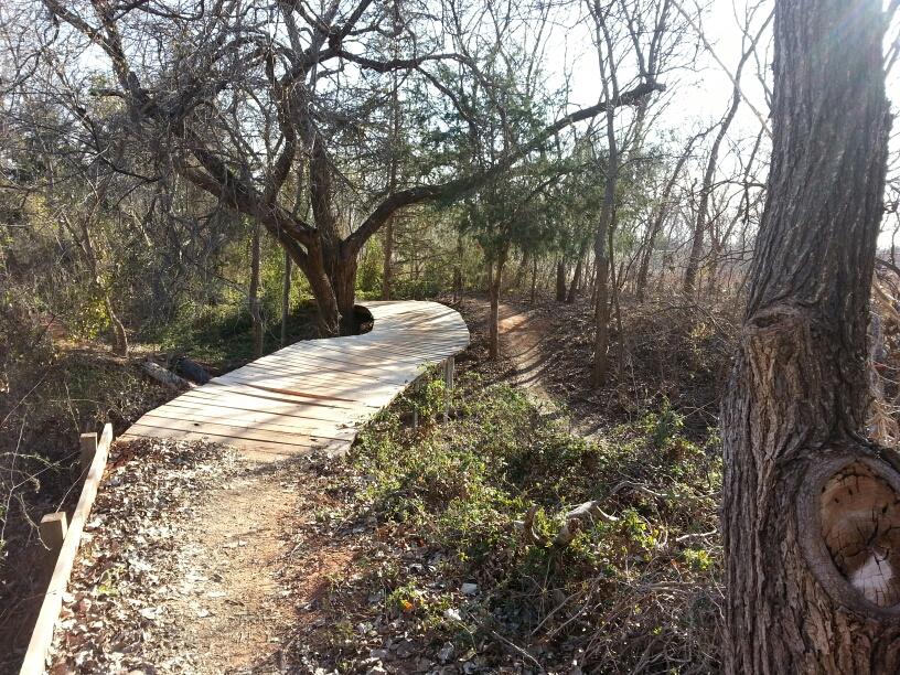 A winding wooden boardwalk surrounded by trees and underbrush, leading through a natural area with sunlight filtering through the branches. Wee-chi-ta mountain bike trail.