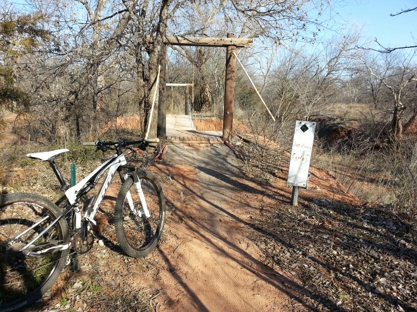 A mountain bike resting on the dirt path near a wooden swing bridge surrounded by bare trees, with a sign indicating the direction to the "Last Area" and "Camping." The scene is set in a natural environment on a clear day. Wee-chi-ta mountain bike trail.