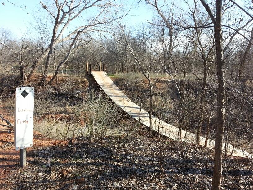 A narrow wooden bridge leading over a small ravine, surrounded by bare trees and sparse underbrush. In the foreground, a sign stands beside the trail, partially obscured by foliage. The scene is bathed in natural light, suggesting a calm, outdoor setting. Wee-chi-ta mountain bike trail.