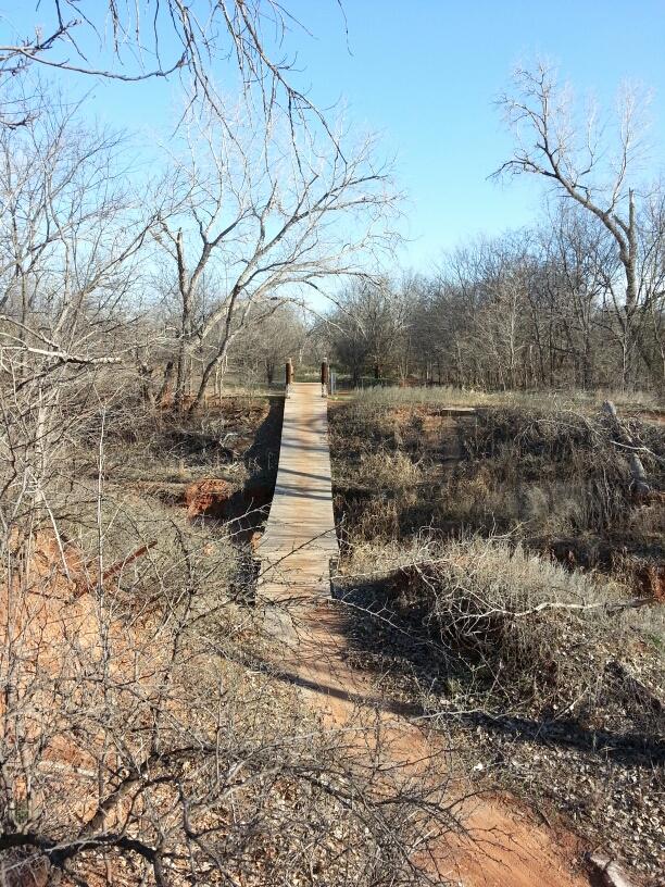 A wooden bridge spanning a dry, rocky area surrounded by sparse trees and brush, under a clear blue sky. The scene evokes a serene and rustic outdoor setting. Wee-chi-ta mountain bike trail.