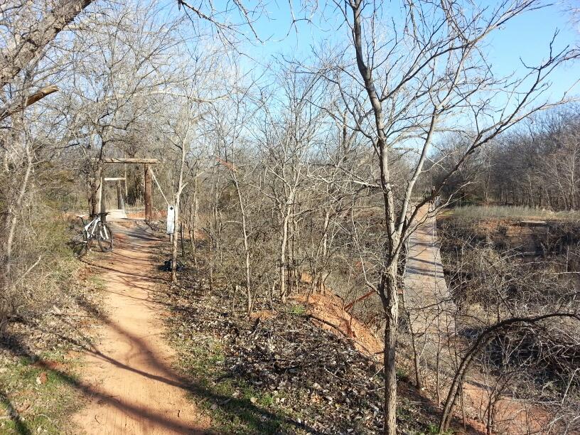 A narrow dirt path winding through a wooded area, lined with bare trees. On the left, there is a bicycle resting against a tree and a simple wooden structure resembling a swing set. In the background, another path leads off into the distance, surrounded by more trees and vegetation under a clear blue sky. Wee-chi-ta mountain bike trail.