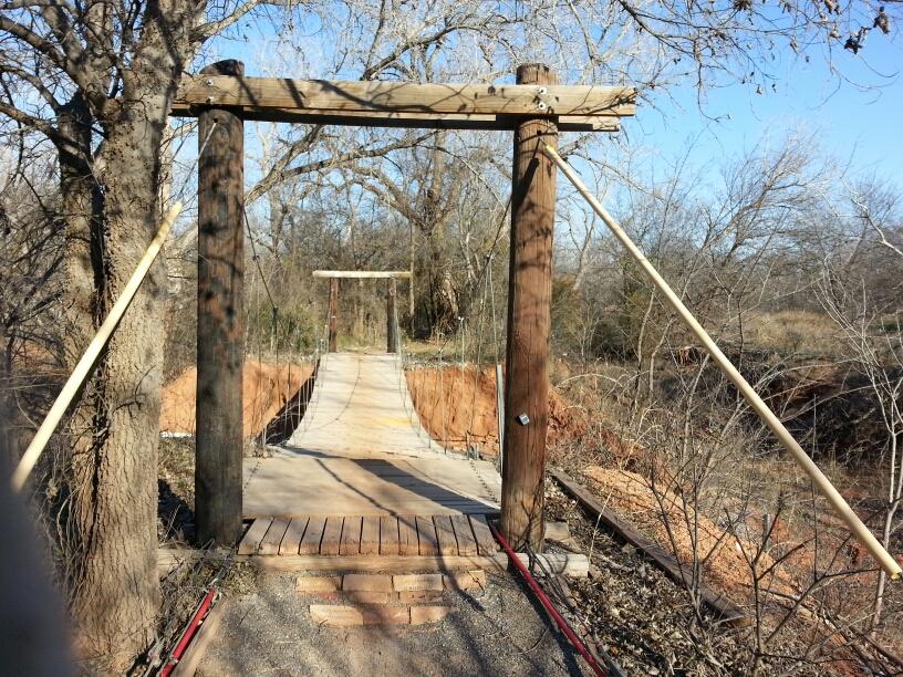 A rustic wooden suspension bridge stretches across a small ravine, supported by sturdy wooden posts on either side. The bridge features a wooden deck and is surrounded by leafless trees and sparse vegetation under a clear blue sky. The scene conveys a natural, outdoor setting. Wee-chi-ta mountain bike trail.