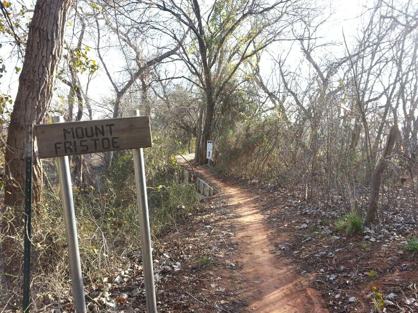 A dirt hiking trail named "Mount Fristoe" is flanked by bare trees and shrubs. A wooden sign marking the trailhead is visible in the foreground, while the path winds into the distance, surrounded by dry leaves and underbrush. The scene is illuminated by sunlight filtering through the trees, creating a tranquil, natural atmosphere. Wee-chi-ta mountain bike trail.