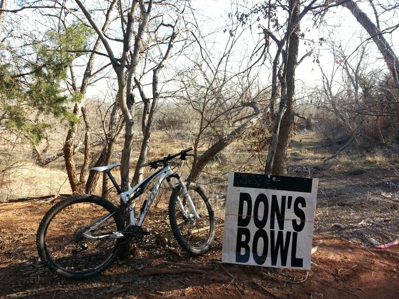 Mountain bike leaning against a sign that reads "DON'S BOWL," surrounded by bare trees and a natural landscape. The scene is set in a wooded area with dirt trail visible in the background. Wee-chi-ta mountain bike trail.