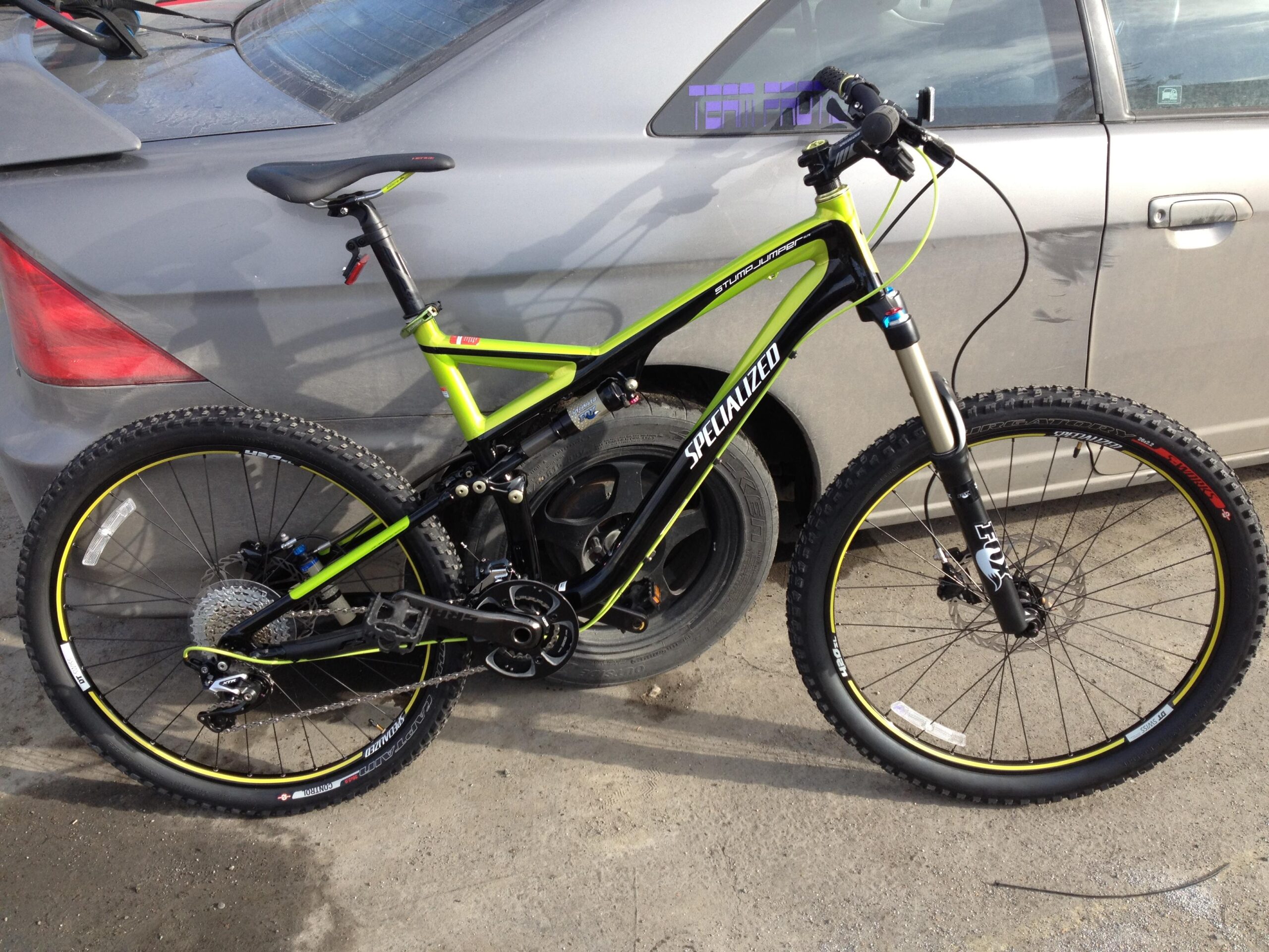 Specialized Stumpjumper FSR Elite: A bright green specialized mountain bike parked next to a gray car. The bike features a dual suspension design, thick tires, and a sleek frame. The background shows part of the car's trunk and a wheel visible in the foreground, with a textured ground surface.