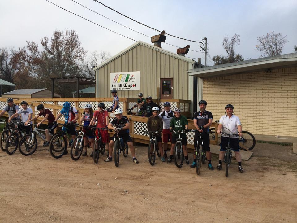 A group of riders, including children and adults, poses with their mountain bikes outside "the BIKE spot," a bike shop. The building has a light-colored exterior, and a wooden deck is visible in front. The riders are wearing helmets and casual clothing, with a mix of bicycles lined up in front of them. The scene is set outdoors with some trees in the background.