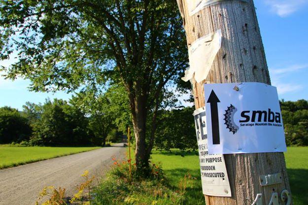 A wooden telephone pole stands beside a gravel road, adorned with multiple signs. One prominent sign featuring the logo of the SMBA (Saratoga Mountain Bike Association) points upward with an arrow, indicating a nearby mountain biking trail. Lush green trees and grass frame the scene, suggesting a natural, outdoor environment. Pittstown State Forest mountain bike trail.
