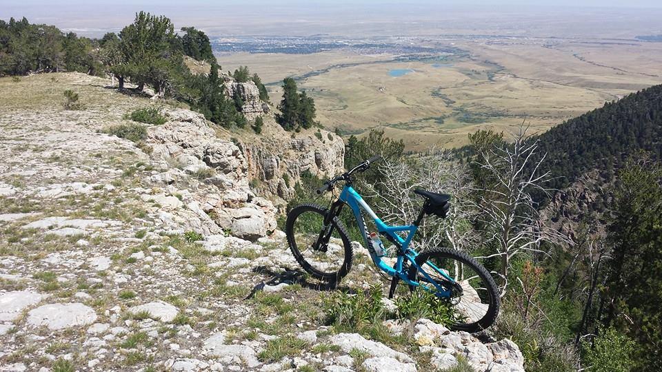 Specialized Stumpjumper FSR Comp EVO: A mountain bike resting on rocky terrain at the edge of a cliff, overlooking a vast valley with greenery and a small lake in the distance under a clear blue sky.