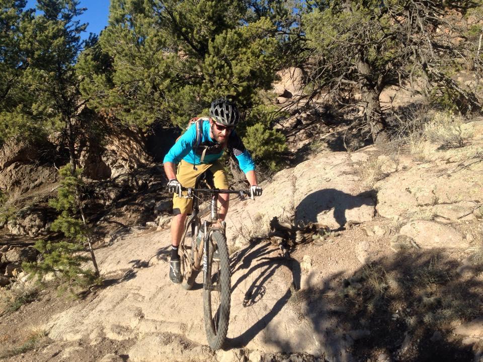 A mountain biker navigates a rocky terrain surrounded by trees, wearing a helmet and sporting a blue and black jersey with khaki shorts. The rider leans forward, focused, as they maneuver over the challenging landscape under clear blue skies. North Backbone mountain bike trail.