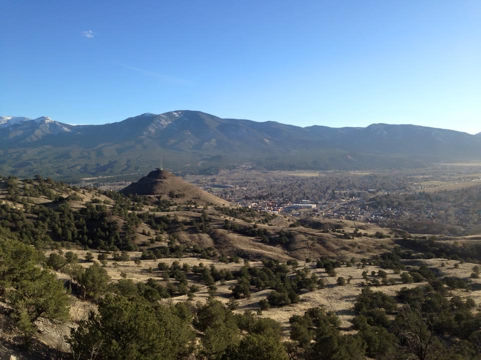 A panoramic view of rolling hills leading up to a mountainous landscape under a clear blue sky. In the foreground, a small, conical hill is visible, surrounded by patches of greenery. In the distance, a town can be seen nestled in the valley, with the surrounding mountains displaying a mix of green and snowy peaks. North Backbone mountain bike trail.