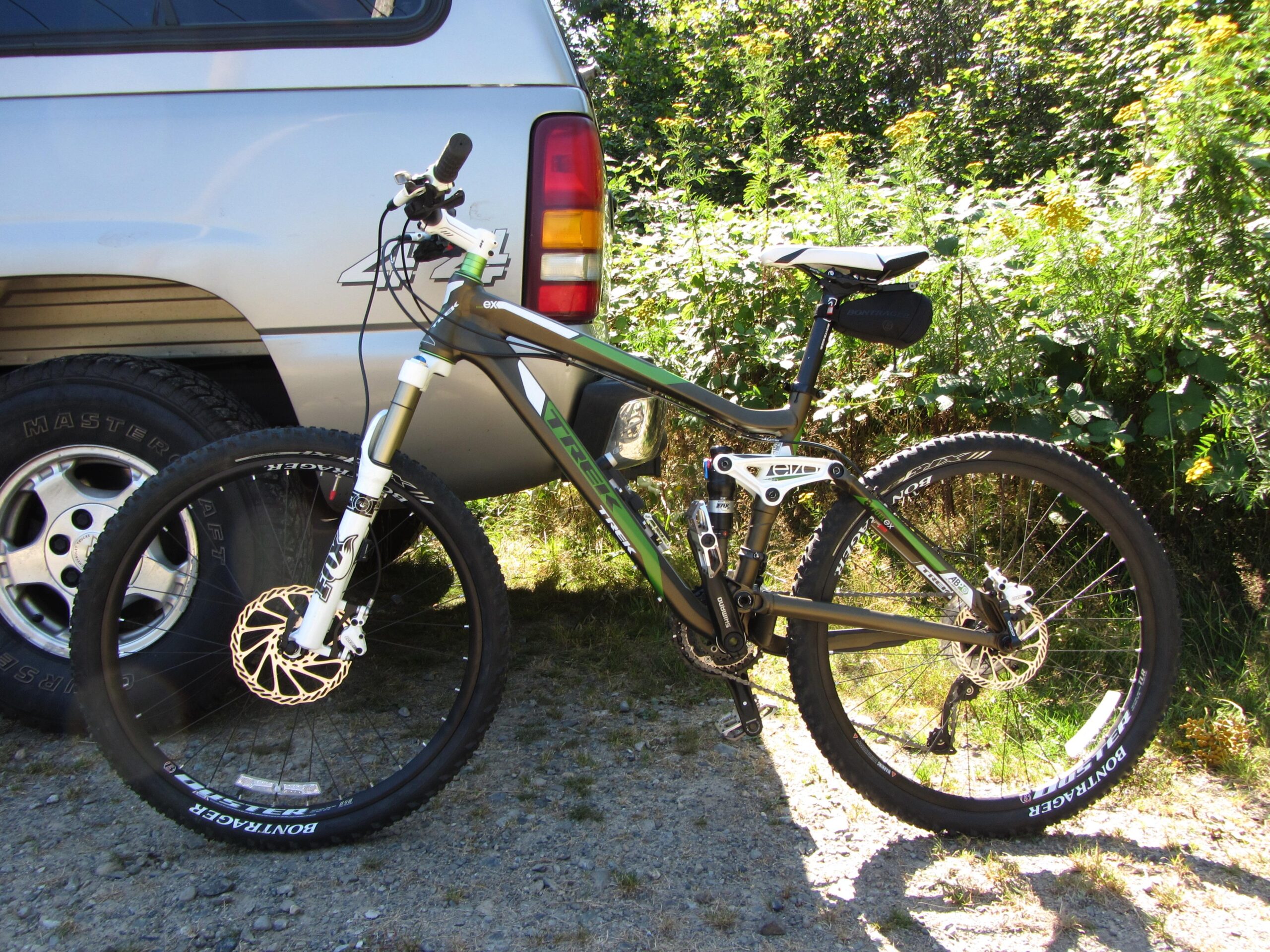 Trek Fuel EX 8: A mountain bike leaning against a silver vehicle on a gravel surface, surrounded by green foliage and sunlight. The bike features a lightweight frame, front suspension, and rugged tires, indicating it's designed for off-road use.