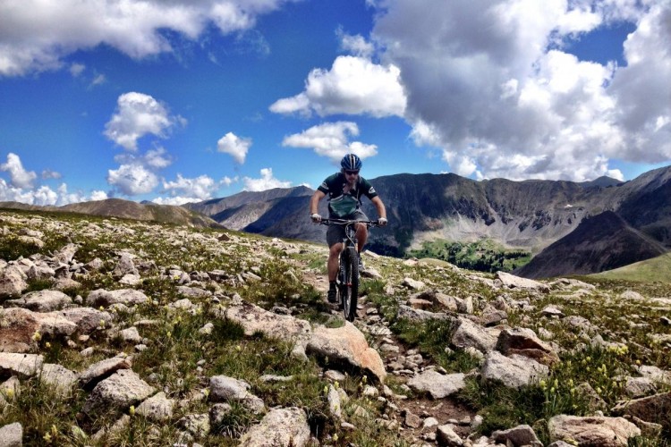 A mountain biker navigates a rocky trail surrounded by grassy terrain and wildflowers, under a bright blue sky filled with fluffy white clouds and rugged mountains in the background.