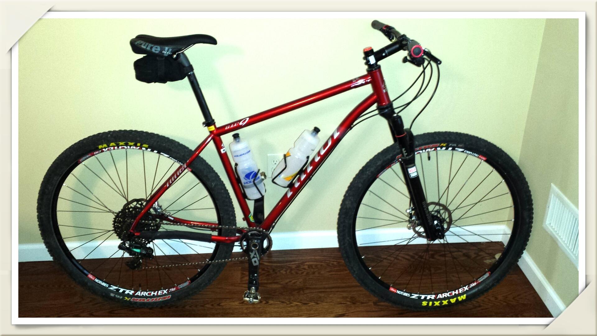 Niner S.I.R. 9: A red mountain bike is positioned against a light-colored wall. The bike features thick tires with visible tread patterns, a black seat, and two water bottles mounted on the frame. The wooden floor adds a warm contrast to the bike's vibrant color.