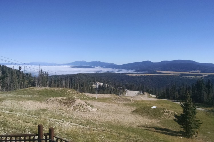 A panoramic view of a mountainous landscape under a clear blue sky. In the foreground, a grassy area is visible, bordered by tall trees. Below, a layer of mist blankets the valley, while mountains rise in the background, creating a serene and breathtaking natural scene.