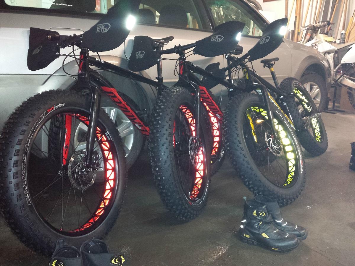 Specialized Fatboy Expert: Three fat bikes with distinctive black frames and colorful wheel accents are parked in a garage. The bikes feature large, textured tires, and each has handlebar mitts. A pair of black and yellow cycling boots sits on the floor beside the bikes, and a vehicle is partially visible in the background.