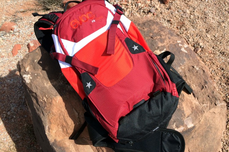 A red and black Evoc FR hiking backpack is resting on a rocky surface. The backpack features a mix of red fabric with white and black accents, showcasing adjustable straps and a zippered pocket. The background consists of gravel and sparse vegetation, indicating an outdoor setting.