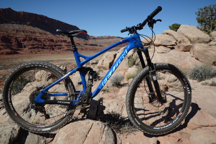 A blue mountain bike parked on rocky terrain with a vast desert landscape in the background, featuring red rock formations under a clear blue sky.