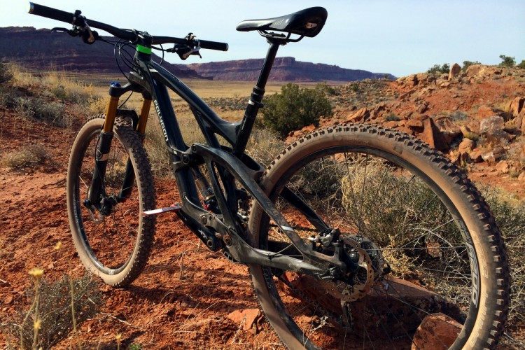 A mountain bike leaning against a rocky terrain, with a background of expansive red desert landscape and distant hills under a clear blue sky. The bike features a black frame, thick tires, and front suspension. Sparse vegetation is visible around the bike, highlighting the rugged outdoor environment.