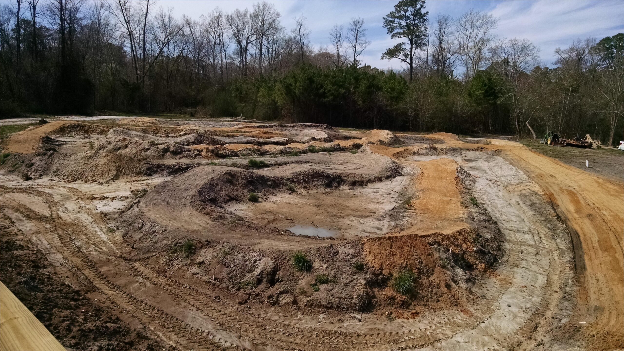 A cleared and partly developed dirt track area with various dirt mounds and shallow depressions, surrounded by sparse trees and brush. The scene features tire tracks marking the ground, indicating prior use, and a small puddle of water in one of the dips. Brown's Creek Nature Park mountain bike trail.