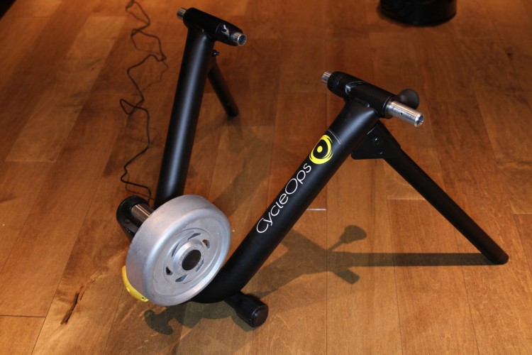 A black CycleOps bike trainer with a silver resistance wheel, positioned on a wooden floor. The trainer features two legs for support and a cable running alongside it.
