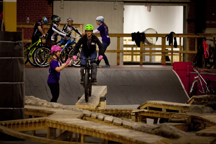 A group of people are participating in a mountain biking lesson indoors. In the foreground, a rider wearing a green helmet is balancing on a wooden ramp, while an instructor in a pink helmet offers support. In the background, several other riders in various helmets are practicing on their bikes, with wooden structures and ramps surrounding them. The environment appears to be a bike park designed for training and skill development.