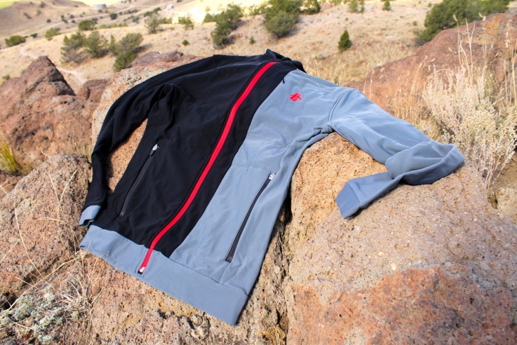 A two-toned athletic jacket lying on a rock in an outdoor setting. The jacket features a black upper section and a gray lower section, with a bright red zipper running down the front and a small logo on the chest. The background shows a natural landscape with trees and hills.