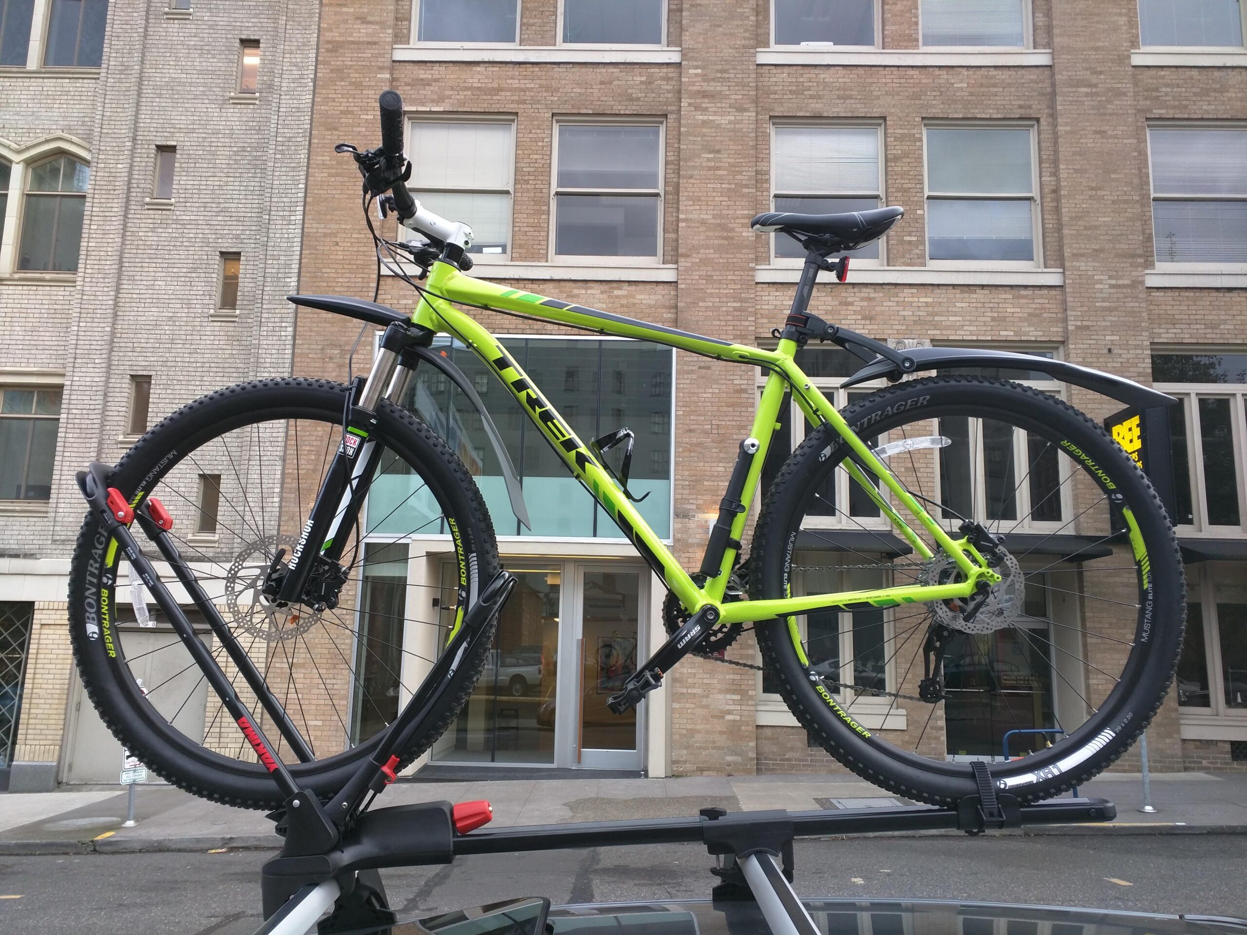 Trek X-Caliber 8: A bright green mountain bike mounted on the roof of a car, with a blurred building facade in the background. The bike features thick tires and a sleek design, showcasing its sporty appearance.