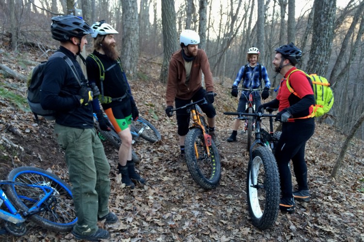 Group of five mountain bikers gathered on a forest trail, surrounded by trees and fallen leaves. They are engaged in conversation, wearing various biking gear, including helmets and colorful jerseys. Bikes are leaning against a nearby tree. The scene conveys a casual outdoor atmosphere during a biking adventure.