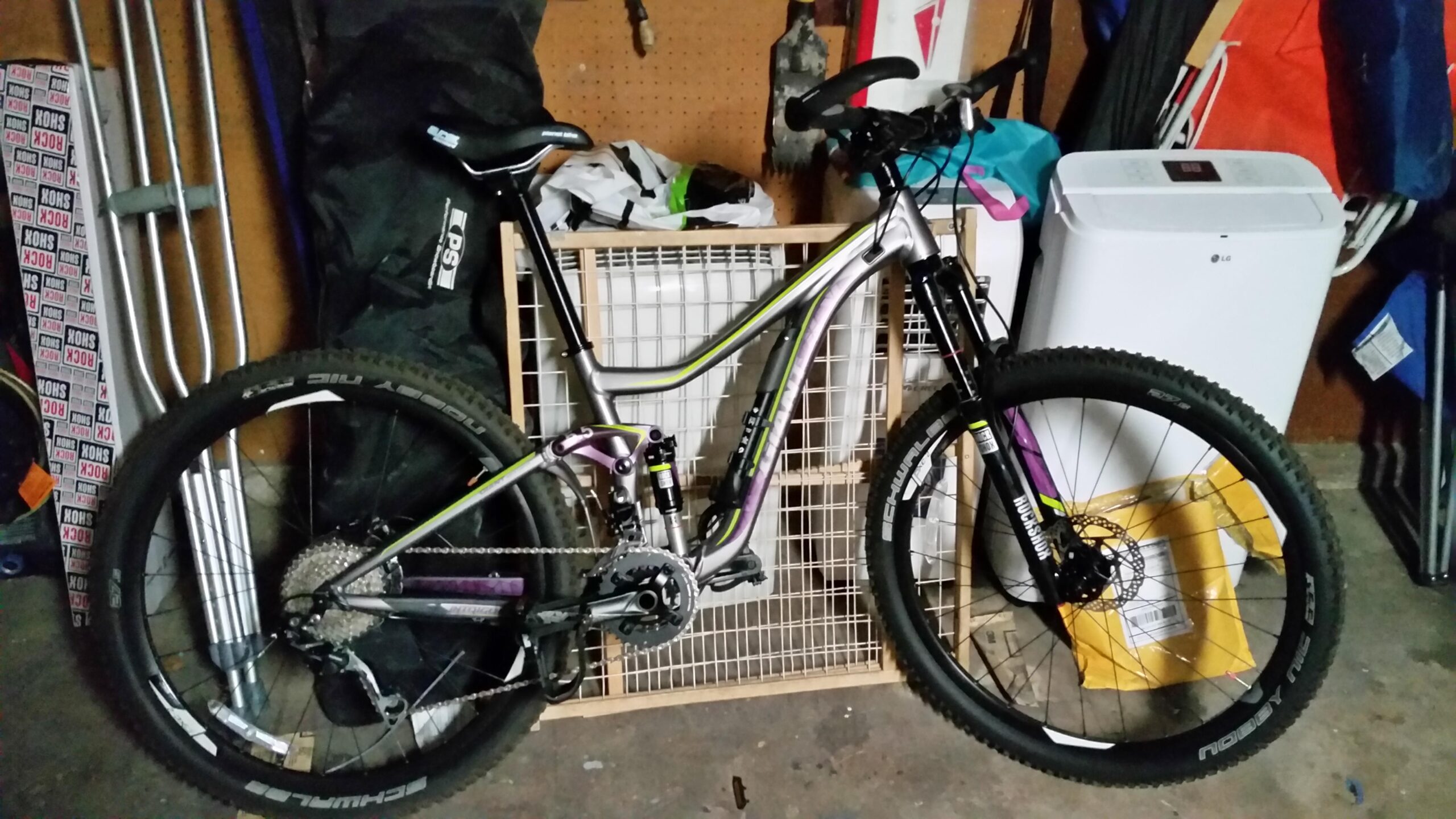 Giant INTRIGUE 2: A mountain bike with a silver and black frame is leaning against a wooden grid in a garage. The bike features thick tires and prominent gears, with various items like crutches and bags visible in the background.