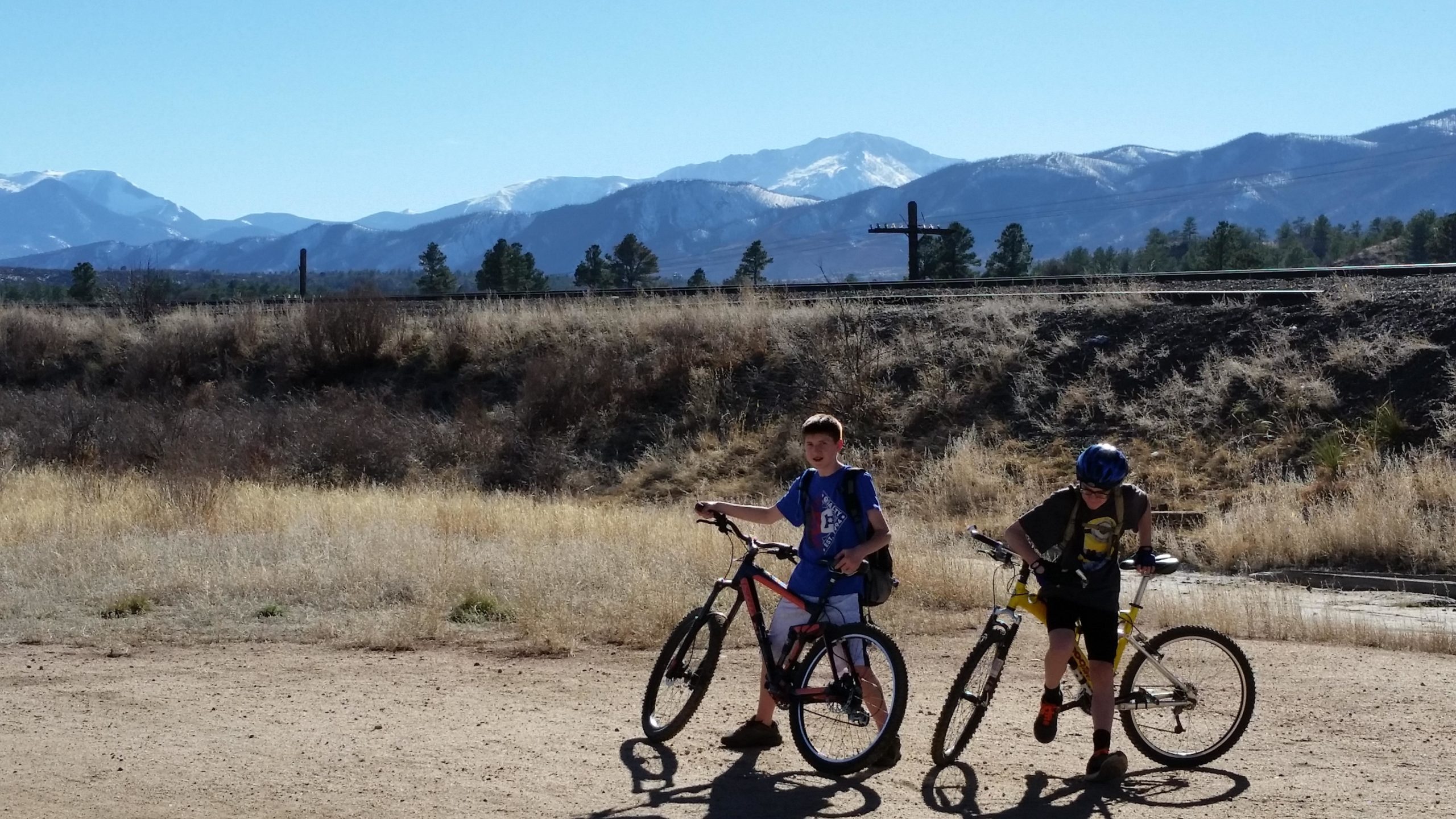 Two boys stand next to their bicycles on a dirt path, with a backdrop of mountains and a clear blue sky. One boy, dressed in a blue shirt and shorts, is holding his bike, while the other boy, wearing a helmet and a gray shirt, is preparing to ride. Surrounding them are dry grasses and shrubs. Santa Fe Trail mountain bike trail.