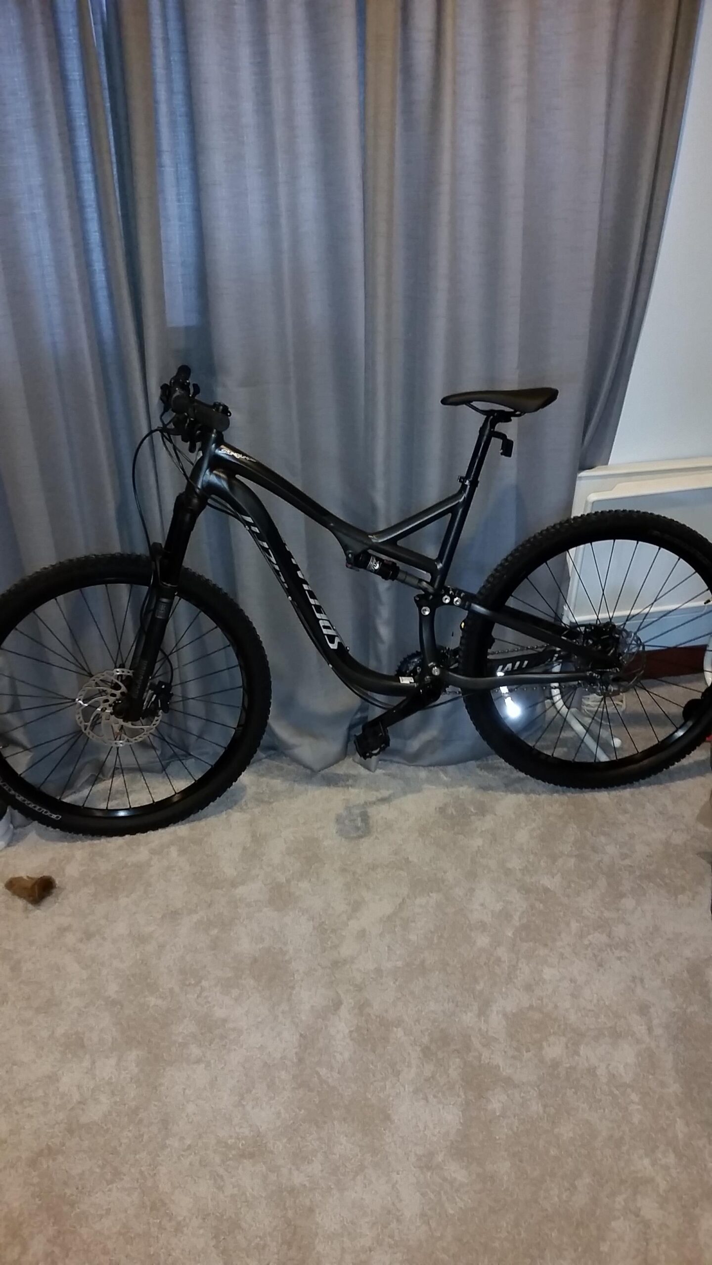 Specialized Stumpjumper FSR Comp 29er: A black mountain bike with thick tires and front suspension stands on a light carpet next to gray curtains. The bike features a sleek frame design and is positioned in a living space.