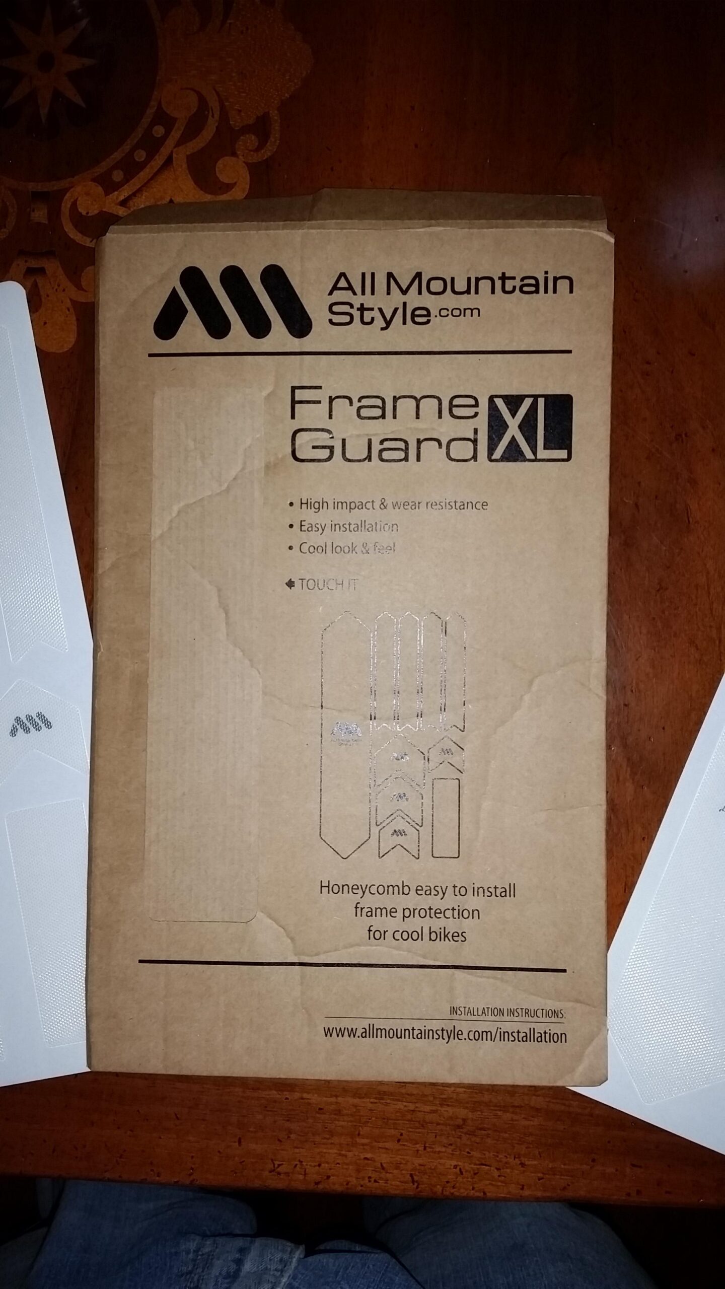 Motobecane NightTrain Bullet: Brown packaging for All Mountain Style Frame Guard XL, featuring text highlighting features such as high impact and wear resistance, easy installation, and a cool look. The package includes images of the frame protection pieces and a website for installation instructions. The background includes a wooden surface.