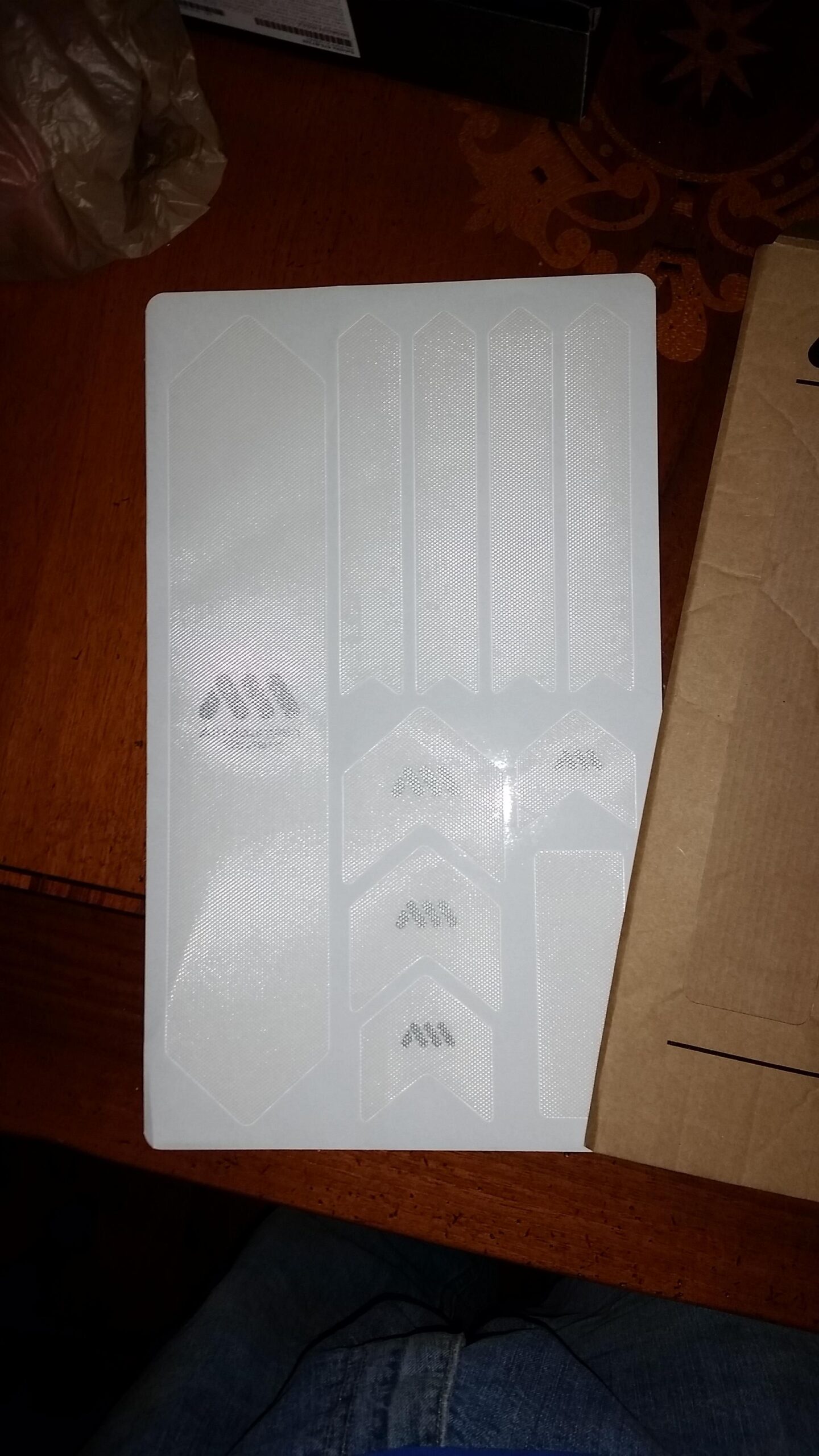 Motobecane NightTrain Bullet: A sheet of white adhesive labels featuring various sizes and shapes, placed on a wooden table. Some of the labels have a textured surface, and the sheet is partially surrounded by other objects, including a plastic bag and a brown cardboard box.